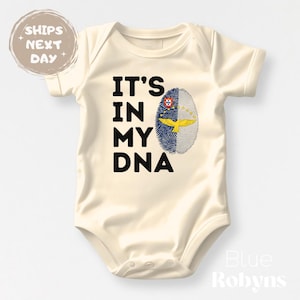 May include: Cream-colored baby bodysuit with the text "IT'S IN MY DNA" in black. A fingerprint design features the Portuguese flag. The bodysuit is a one-piece garment.