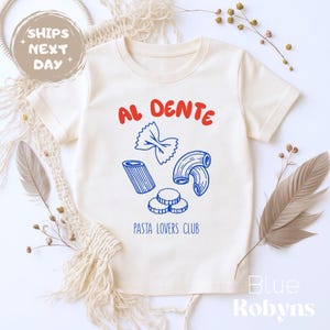 May include: Cream-colored t-shirt with the words "AL DENTE" in red above blue pasta illustrations. Below the pasta, the text "PASTA LOVERS CLUB" is printed in blue. The shirt has short sleeves and a crew neck.