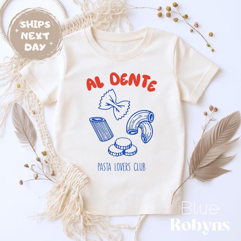 May include: Cream-colored t-shirt with the words "AL DENTE" in red above blue pasta illustrations. Below the pasta, the text "PASTA LOVERS CLUB" is printed in blue. The shirt has short sleeves and a crew neck.