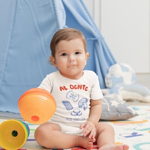 May include: A baby wearing a white onesie with the words "AL DENTE" in red and blue, along with pasta illustrations. The baby is holding an orange ball and sitting on a colorful play mat. A blue teepee is in the background.