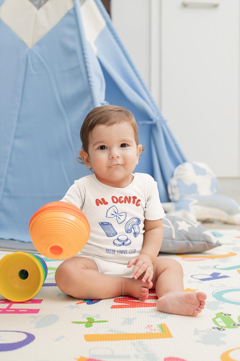 May include: A baby wearing a white onesie with the words "AL DENTE" in red and blue, along with pasta illustrations. The baby is holding an orange ball and sitting on a colorful play mat. A blue teepee is in the background.