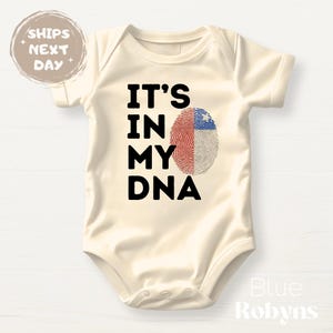 May include: Cream-colored baby bodysuit with the text "IT'S IN MY DNA" in black. The design includes a fingerprint split with red, white, and blue colors, representing a flag. The bodysuit is a one-piece garment.