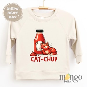 May include: A cream colored sweatshirt with a red cat and a ketchup bottle. The text "CAT-CHUP" is printed on the shirt.