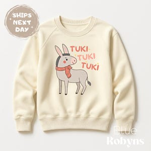 May include: Cream-colored sweatshirt featuring a cartoon donkey wearing a red scarf. The donkey has gray fur and is positioned in the center of the sweatshirt. The words "TUKI TUKI TUKI" are printed in pink above the donkey.
