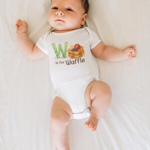 May include: A white baby onesie with the text "W is for Waffle" and a watercolor illustration of waffles, blueberries, and a strawberry. The baby is lying on a white sheet.