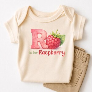 May include: A cream-colored baby onesie with a pink letter "R" and a watercolor raspberry illustration. The text "R is for Raspberry" is below the letter and fruit. Tan pants are folded next to the onesie.