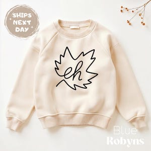 May include: Cream-colored sweatshirt with a black maple leaf design and the word "eh" inside. The sweatshirt has a crew neck and long sleeves. The text "Blue Robyn's" is at the bottom right.