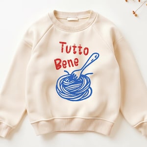 May include: Cream-coloured sweatshirt with a crew neck and raglan sleeves. The front features the Italian phrase "Tutto Bene" in red, above a blue illustration of spaghetti and a fork.