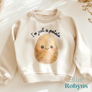 May include: Cream-colored sweatshirt featuring a smiling potato graphic with the text "I'm just a potato." The potato has rosy cheeks and a happy expression. The sweatshirt has a ribbed neckline, cuffs, and hem.
