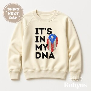Puede incluir: Sudadera color crema con el texto "IT'S IN MY DNA" en negro. Un dise&ntilde;o de huella dactilar con los colores de la bandera de Puerto Rico est&aacute; a la derecha. La sudadera tiene cuello redondo y mangas largas. La marca "Blue Robyn's" est&aacute; en la esquina inferior derecha.