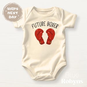 May include: Cream-colored baby onesie with the text "FUTURE BOXER" above a graphic of two red boxing gloves. The onesie is a short-sleeved garment, suitable for infants.