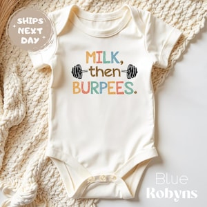 May include: Cream-colored baby onesie with the text "MILK, then BURPEES" in colorful letters and a barbell graphic. The onesie has short sleeves and snaps at the bottom. The text "Blue Robyn's" is in the bottom right corner.