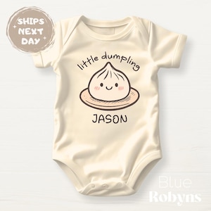 May include: Cream-colored baby onesie with the text "little dumpling" above a cartoon dumpling illustration. The name "JASON" is printed below the dumpling. The onesie has short sleeves and a round neckline.