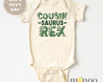 May include: A cream-colored baby bodysuit with short sleeves and a green dinosaur-themed graphic that says "COUSIN-SAURUS-REX".