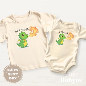 May include: Two matching cream-colored shirts with dragon graphics. One is a t-shirt with "Big Dragon" text and a green dragon breathing fire. The other is a baby bodysuit with "Lil Dragon" text and a similar dragon design.