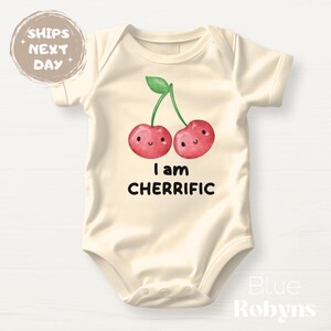 May include: Cream-colored baby bodysuit with a cheerful cherry design. The cherries are red with smiling faces and a green stem, and the text "I am CHERRIFIC" is printed below. A cute and playful outfit for infants.
