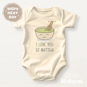 May include: A cream-colored baby onesie with a cartoon illustration of a bowl of green matcha with a whisk and the text "I love you so matcha".