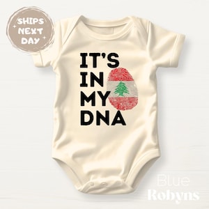 May include: Cream-colored baby bodysuit with the text "IT'S IN MY DNA" in black. A fingerprint design in red and green, representing the Lebanese flag, is centered on the front. The bodysuit has short sleeves.