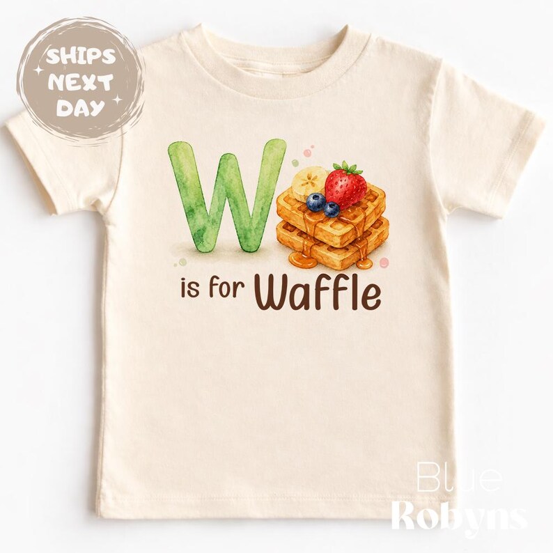 May include: Cream-colored t-shirt featuring a watercolor design. A large green "W" is next to a stack of waffles topped with fruit and syrup. The text "is for Waffle" is below the image. The shirt has short sleeves.