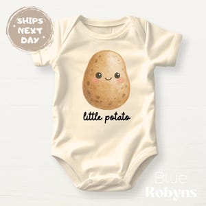 May include: Cream-colored baby bodysuit featuring a smiling potato graphic with the text "little potato" in black. The potato has rosy cheeks and a cute expression. The bodysuit is short-sleeved and made of soft material.