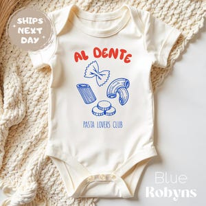 May include: Cream-colored baby onesie with a pasta-themed graphic. The design includes the words "AL DENTE" in red, and blue illustrations of different pasta shapes. The text "PASTA LOVERS CLUB" is below the pasta illustrations.
