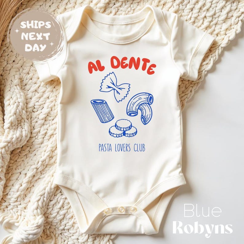 May include: Cream-colored baby onesie with a pasta-themed graphic. The design includes the words "AL DENTE" in red, and blue illustrations of different pasta shapes. The text "PASTA LOVERS CLUB" is below the pasta illustrations.
