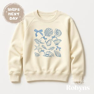 May include: A cream-colored sweatshirt with a blue seashell and bow design. The sweatshirt has a crew neck and long sleeves.