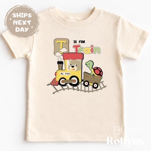 May include: Cream-coloured toddler t-shirt with a colourful train graphic. The train has a bear conductor and a turtle passenger. Text on the shirt reads "T is for Train". The shirt also has the text "Blue Robyns".