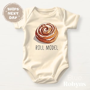 May include: A cream-colored baby onesie with a cinnamon roll illustration and the text "Roll Model".