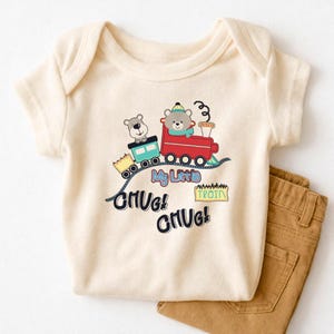 May include: Cream baby bodysuit with a train graphic featuring two bears. The train is red and teal, with the words "My Little Train" and "Chug! Chug!" printed in blue and black. Brown trousers are folded next to the onesie.