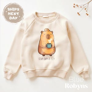 May include: Cream-colored sweatshirt featuring a cartoon capybara holding a blue mug with steam rising. The text "STAY CAPY & COZY" is below the capybara. The sweatshirt has a crew neck and long sleeves.