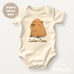 May include: A cream-colored baby onesie with a cartoon capybara wearing a pink and white donut on its head. The onesie has the text "Custom Name" printed on the front.