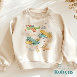 May include: Cream-colored sweatshirt featuring a watercolor design of several airplanes in shades of green, yellow, blue, and orange. The design includes clouds and stars. The text "Blue Robyn's" is printed in teal at the bottom right.
