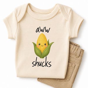 May include: A cream-colored short-sleeve bodysuit featuring a graphic of a smiling yellow corn cob with green husks. The text "aww" is printed above the corn and "shucks" is printed below in a simple black font, paired with tan pants.