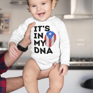 Puede incluir: Un body blanco de beb&eacute; con mangas largas que presenta el texto "IT'S IN MY DNA" y un dise&ntilde;o de huella dactilar con los colores de la bandera de Puerto Rico. El beb&eacute; est&aacute; sonriendo.