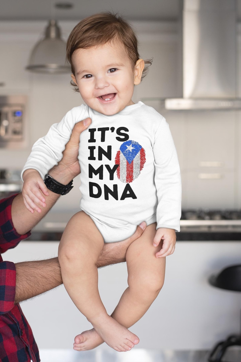 Puede incluir: Un body blanco de beb&eacute; con mangas largas que presenta el texto "IT'S IN MY DNA" y un dise&ntilde;o de huella dactilar con los colores de la bandera de Puerto Rico. El beb&eacute; est&aacute; sonriendo.
