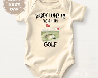May include: Cream-colored baby onesie with the text "DADDY LOVES ME more than GOLF". The design includes a golf hole with a red flag, a golf ball, and a small red heart. The onesie is short-sleeved.