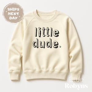 Peut inclure: Sweat-shirt crème avec l'inscription "little dude" en noir, lettres soulignées. Le sweat-shirt a un col rond et des manches raglan. Les mots "Ships Next Day" sont dans un cercle en haut à gauche.