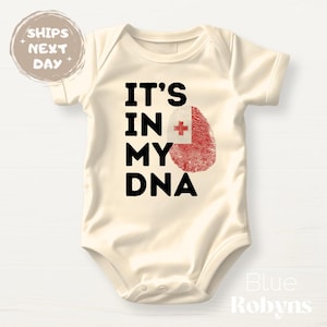 May include: Cream-colored baby bodysuit with the text "IT'S IN MY DNA" in black. A red fingerprint with a white and red cross graphic is next to the text. The words "Blue Robyns" are in the bottom right corner.