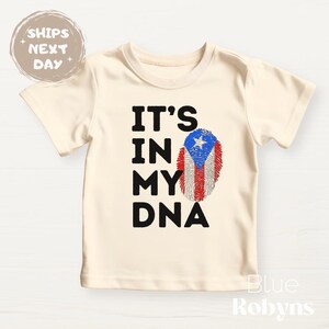 Puede incluir: Camiseta color crema con el texto "IT'S IN MY DNA" en letras negras. Un dise&ntilde;o de huella dactilar con los colores de la bandera de Puerto Rico est&aacute; presente. La camiseta es de manga corta.