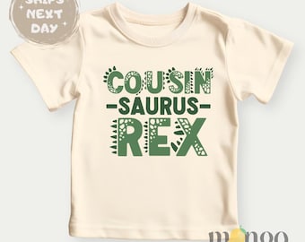 May include: A cream colored t-shirt with green text that reads "Cousin-saurus Rex".
