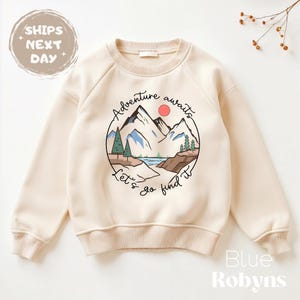 May include: Cream-colored sweatshirt with a circular graphic featuring a mountain landscape, a red sun, and the words "Adventure awaits. Let's go find it." The sweatshirt has long sleeves and a crew neck.