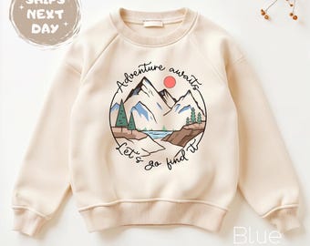 May include: Cream-colored sweatshirt with a circular graphic featuring a mountain landscape, a red sun, and the words "Adventure awaits. Let's go find it." The sweatshirt has long sleeves and a crew neck.