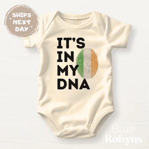 May include: A cream-coloured baby bodysuit featuring the text "IT'S IN MY DNA" in black. The "A" in DNA is replaced with a fingerprint design in the colours of the Irish flag. A round sticker that says "SHIPS NEXT DAY" is in the upper left corner.