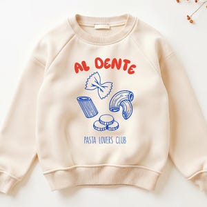 May include: Cream-colored sweatshirt with the words "AL DENTE" in red above blue pasta illustrations. Below the pasta, the text reads "PASTA LOVERS CLUB". The raglan sleeves and crew neck are the same color as the body.