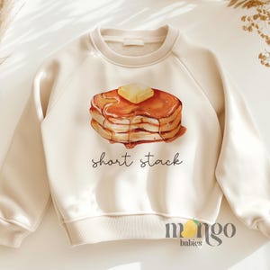 May include: A cream-colored sweatshirt with a graphic of a short stack of pancakes with butter and syrup. The text "short stack" is printed below the pancakes.