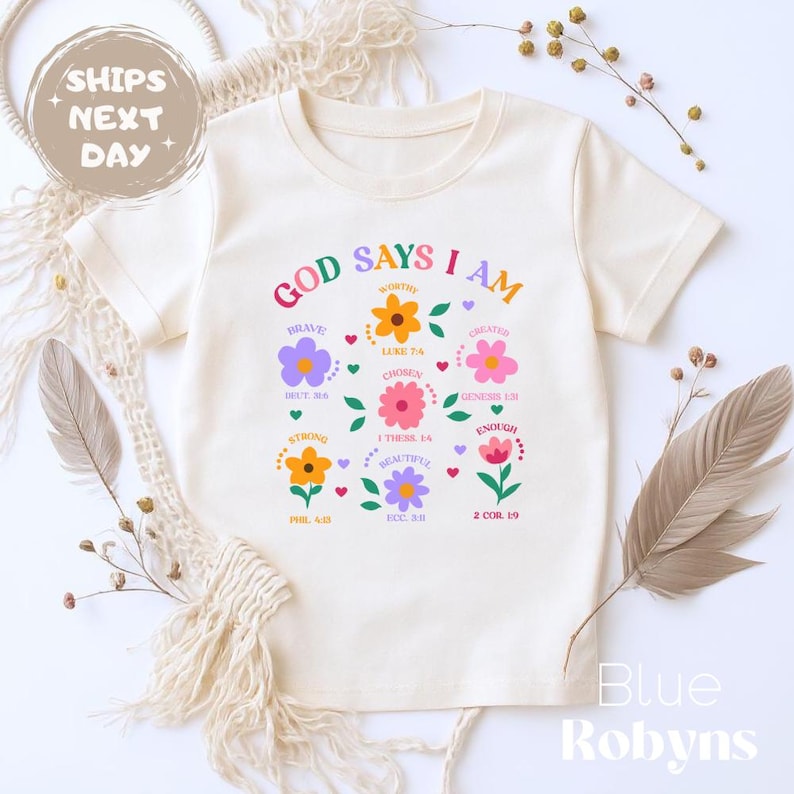 Op de afbeelding: Cr&egrave;mekleurig T-shirt met de tekst "GOD SAYS I AM" en bloemmotieven. Het shirt heeft woorden als "Brave", "Worthy" en "Chosen" in de bloemengraphics. De tekst "Blue Robyns" staat rechtsonder.