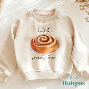 May include: A cream-colored sweatshirt with a graphic of a cinnamon roll and the text "little sweet bun".