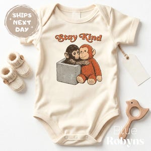 May include: Cream-colored baby onesie with the text "Stay Kind" in orange script. The onesie features a graphic of two cartoon monkeys hugging. A pair of knitted baby booties and a wooden bird-shaped toy are also visible.