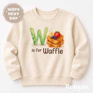 May include: Cream-colored sweatshirt with a watercolor design. The design features a large green letter "W", the phrase "is for Waffle", and a stack of waffles topped with fruit. The sweatshirt is a crew neck style.